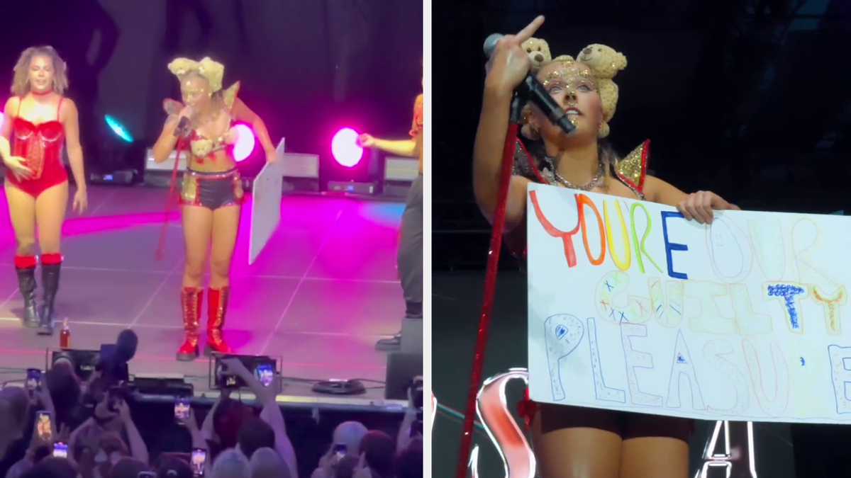 Jojo Siwa on stage holding a sign that reads "YOU'RE OUR GUILTY PLEASURE," while performing alongside another dancer in a red outfit
