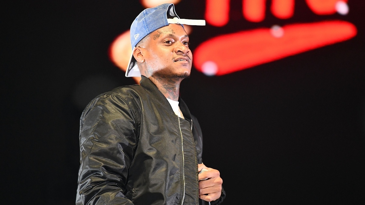 Young man wearing a baseball cap backward and a bomber jacket on stage. Text in the background is blurred