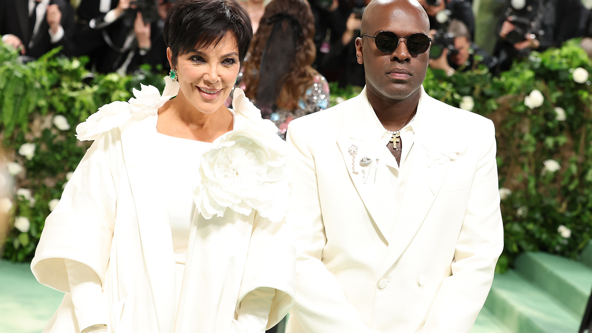 Kris Jenner and Corey Gamble at a formal event. Kris is in an elegant white outfit with large flower details, while Corey wears a sleek white suit and sunglasses
