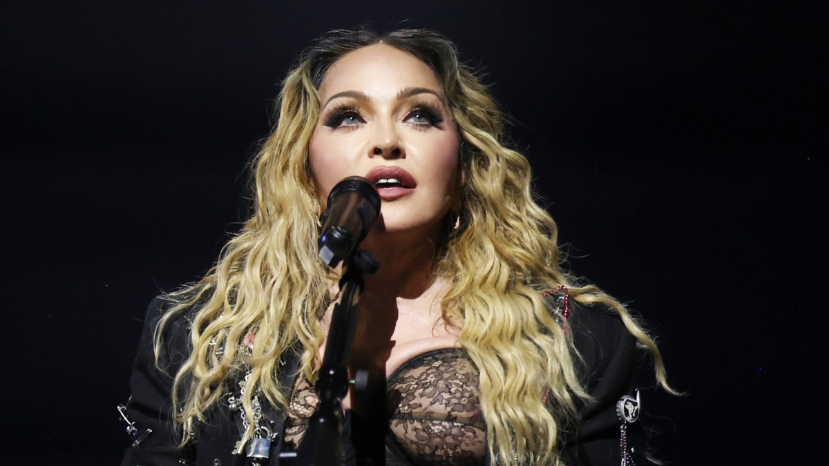 Madonna performs on stage, holding a microphone. She has wavy blonde hair and is wearing a black lace top under a black jacket