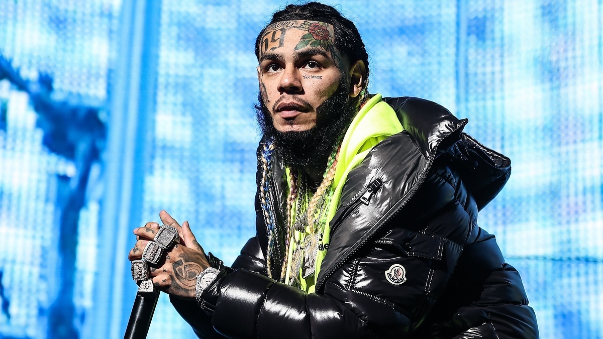 Tekashi 6ix9ine, with face tattoos, a heavy jacket, and multiple rings with "69" inscription, performing on stage, leaning forward while holding a microphone