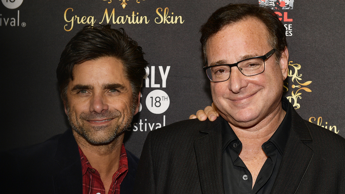 John Stamos and Bob Saget pose together at an event. John wears a checkered shirt and casual jacket, while Bob is in a dark suit with a collared shirt