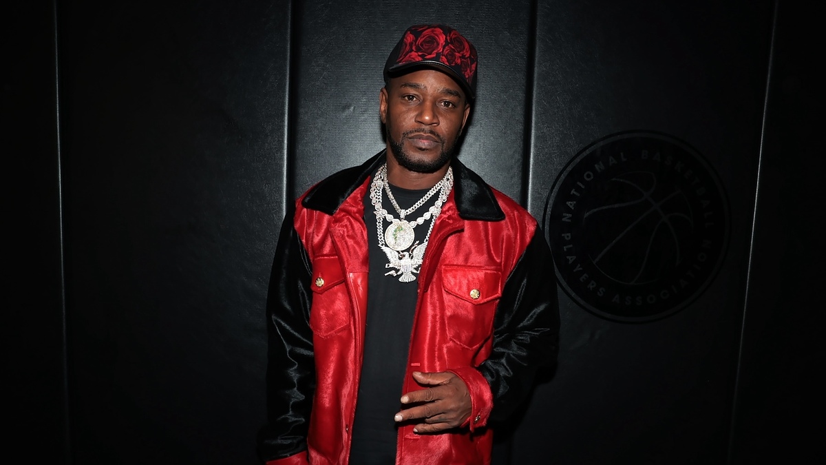 A man stands against a dark backdrop wearing a red and black jacket, a red hat, and layered silver chains