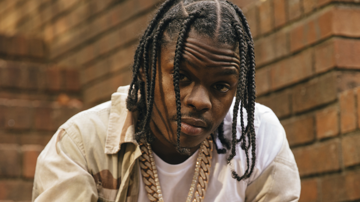 A man is sitting on brick steps, wearing a white shirt, a tan jacket, and pants, accessorized with gold chains and a gold watch
