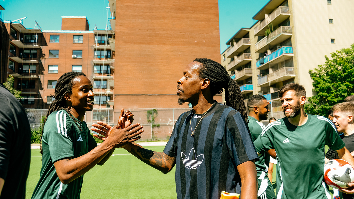 Two soccer players, one in a green jersey and another in a black Adidas shirt, greeting each other on a soccer field with teammates in the background