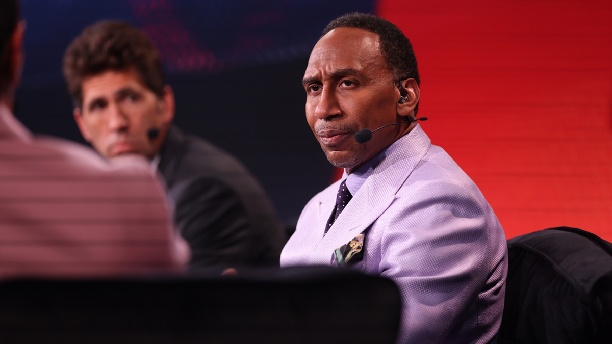 Two people sitting on a panel with microphones. The man in front wears a suit and tie; the man behind him also wears a suit