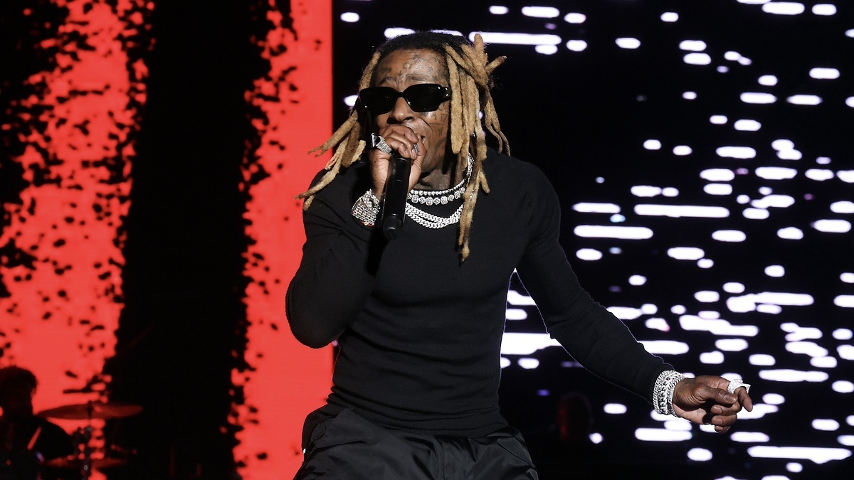 Lil Wayne performs on stage, wearing a black long-sleeve shirt, multiple chains, and sunglasses, with colorful lights in the background