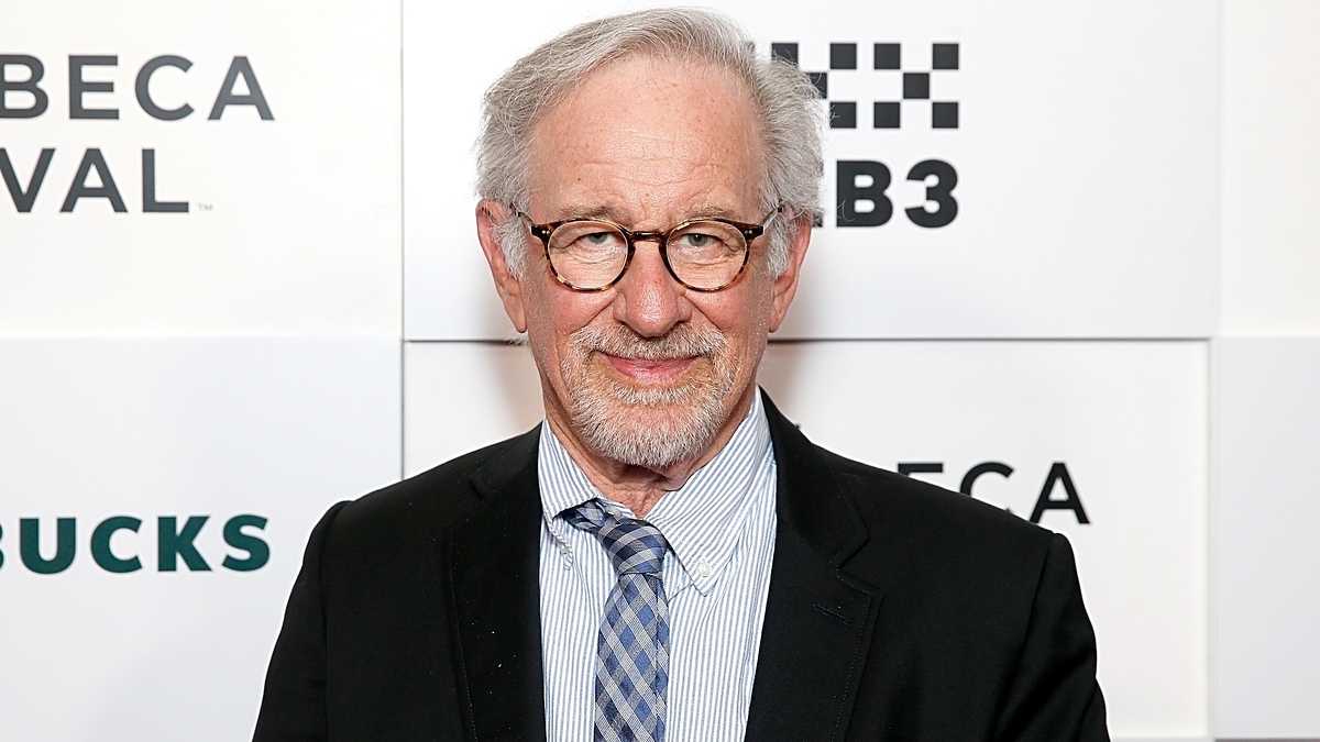 Steven Spielberg at an event, wearing a suit with a striped shirt and patterned tie, in front of a backdrop with logos including Tribeca Festival and Starbucks