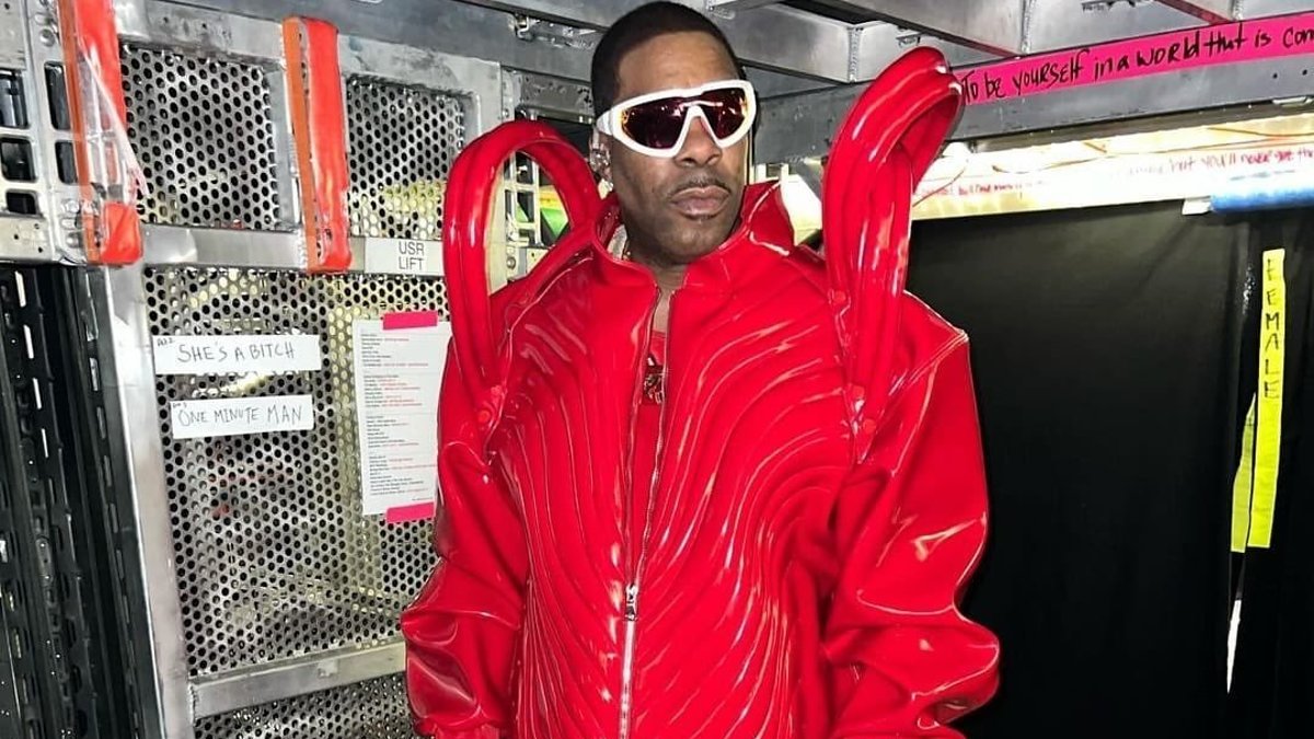 A person wearing an avant-garde, red outfit with exaggerated shoulders and flared pants, paired with white sunglasses, posing backstage