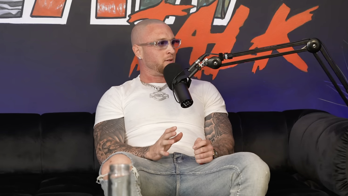 A bald man with tattoos, wearing a white t-shirt and jeans, speaks into a microphone while seated on a black couch, on a podcast set with vibrant wall art