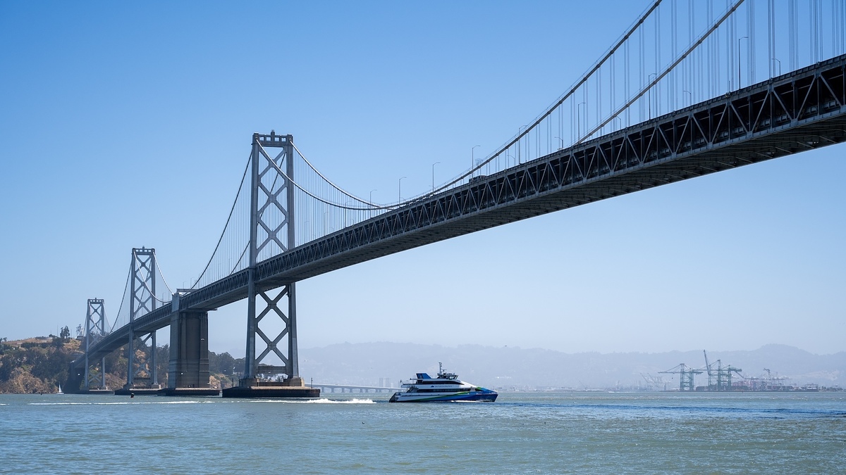 San Francisco Bay Bridge with a ferry boat crossing beneath, mountains and city skyline in the background