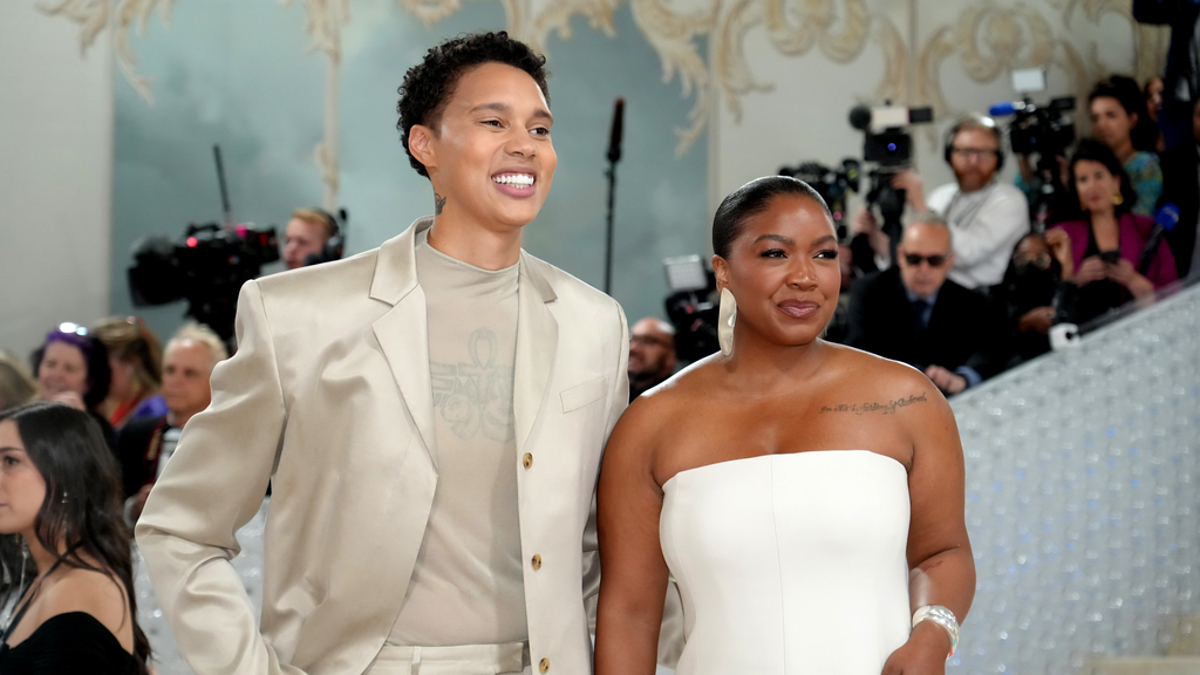 Brittney Griner in a light suit and Cherelle Griner in a strapless dress pose together at an event with photographers in the background