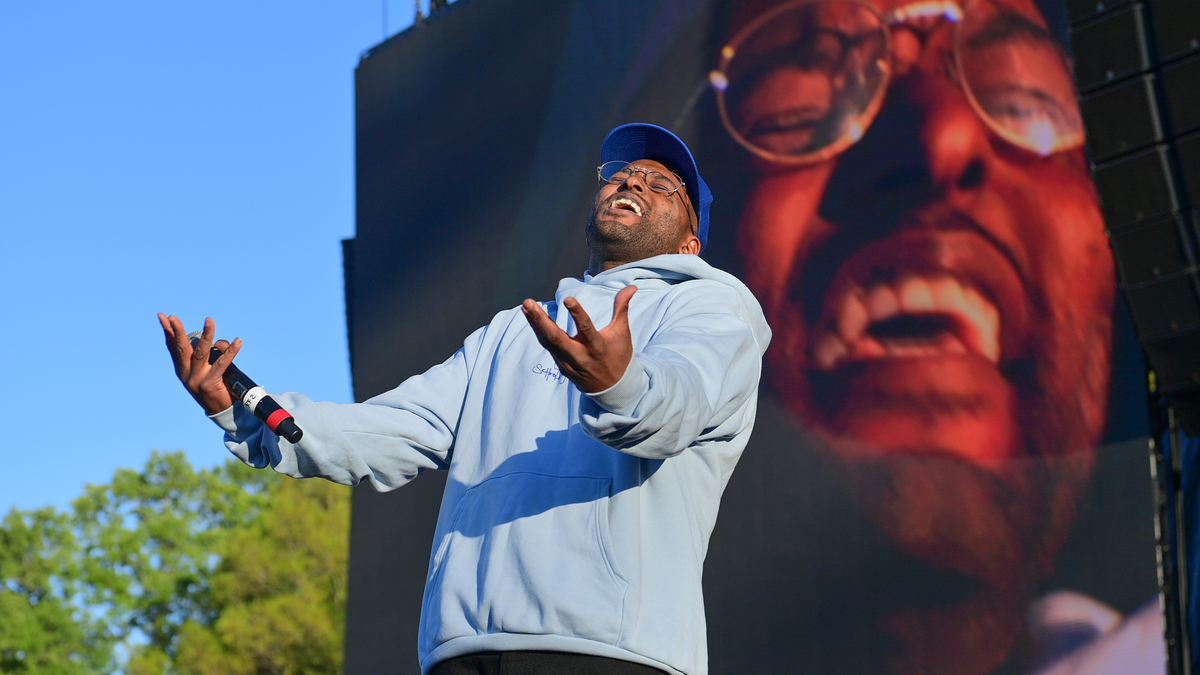 Man on stage performing, wearing a light hoodie and cap, with his face displayed prominently on a large screen behind him during a music event