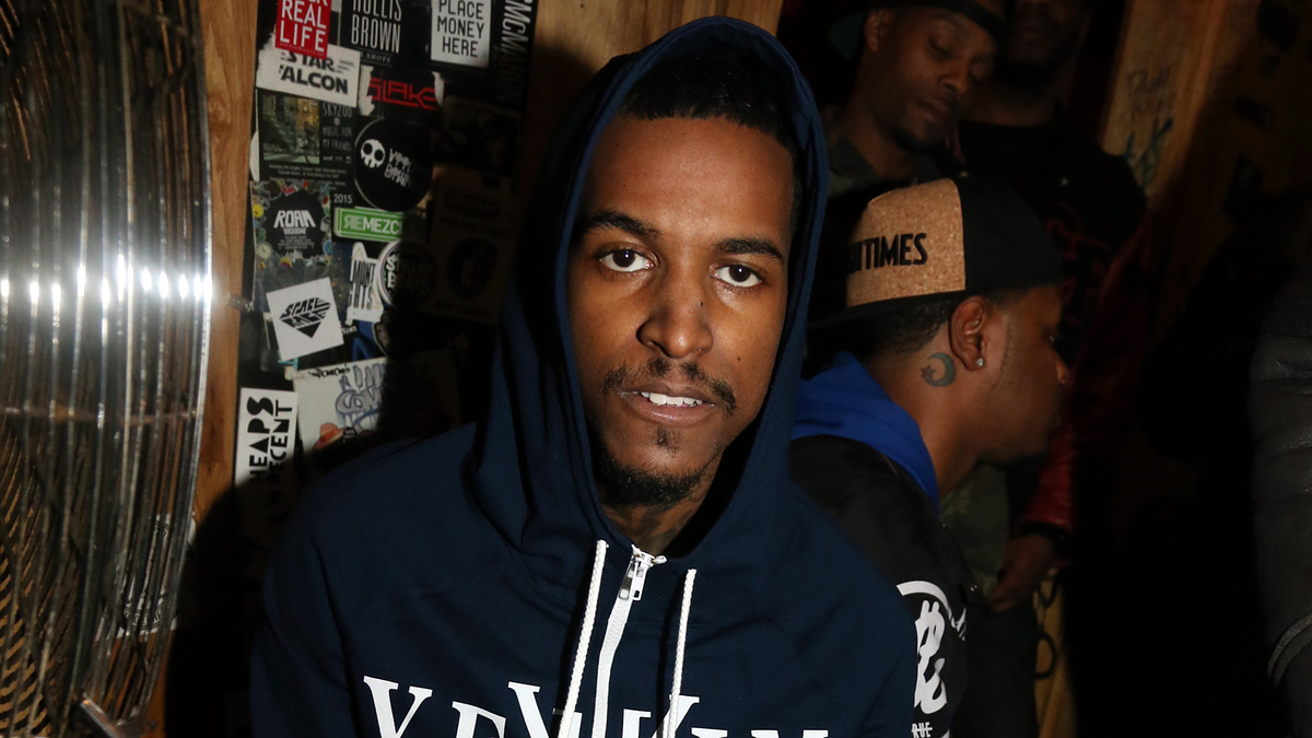 A man wearing a hoodie stands in front of a wall covered with stickers and graffiti, with others in the background. The image is categorized under Music