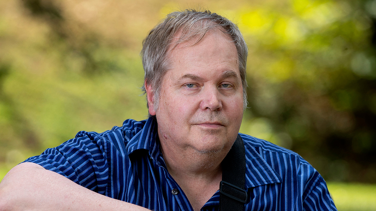 John Hinckley Jr. sits outdoors on a bench, holding a guitar inscribed with his name, wearing a striped shirt. The background is blurred greenery