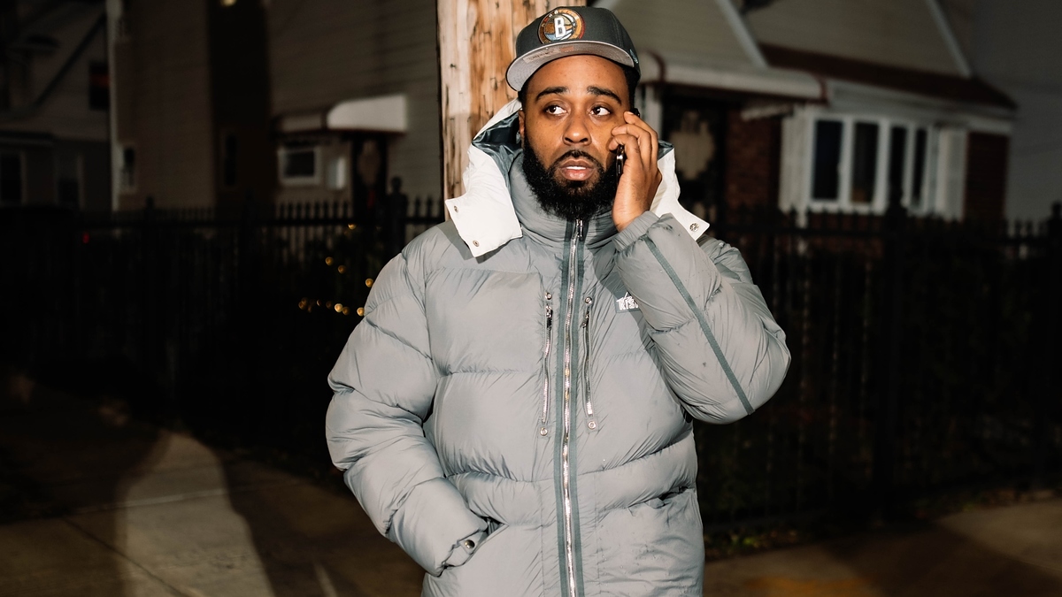 Man wearing a thick winter jacket and cap talking on the phone while standing outdoors on a residential street at night