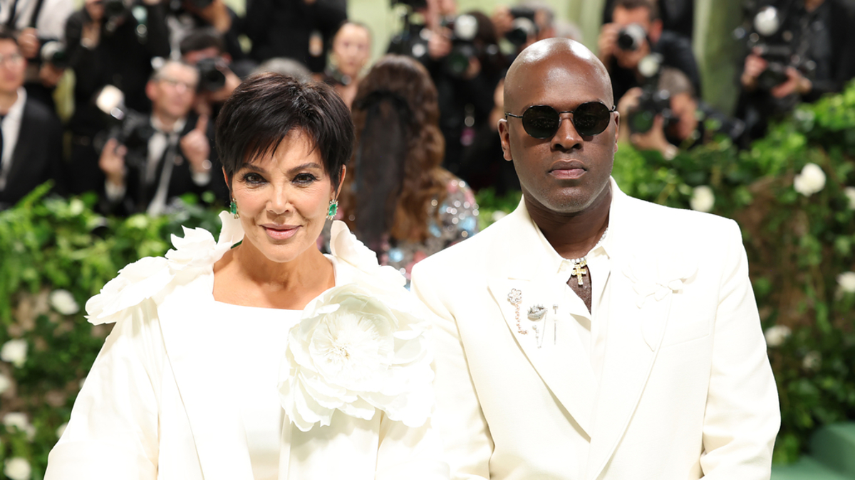 Kris Jenner and Corey Gamble attend an event, both wearing white outfits with floral accents. Kris has short hair, and Corey is wearing sunglasses