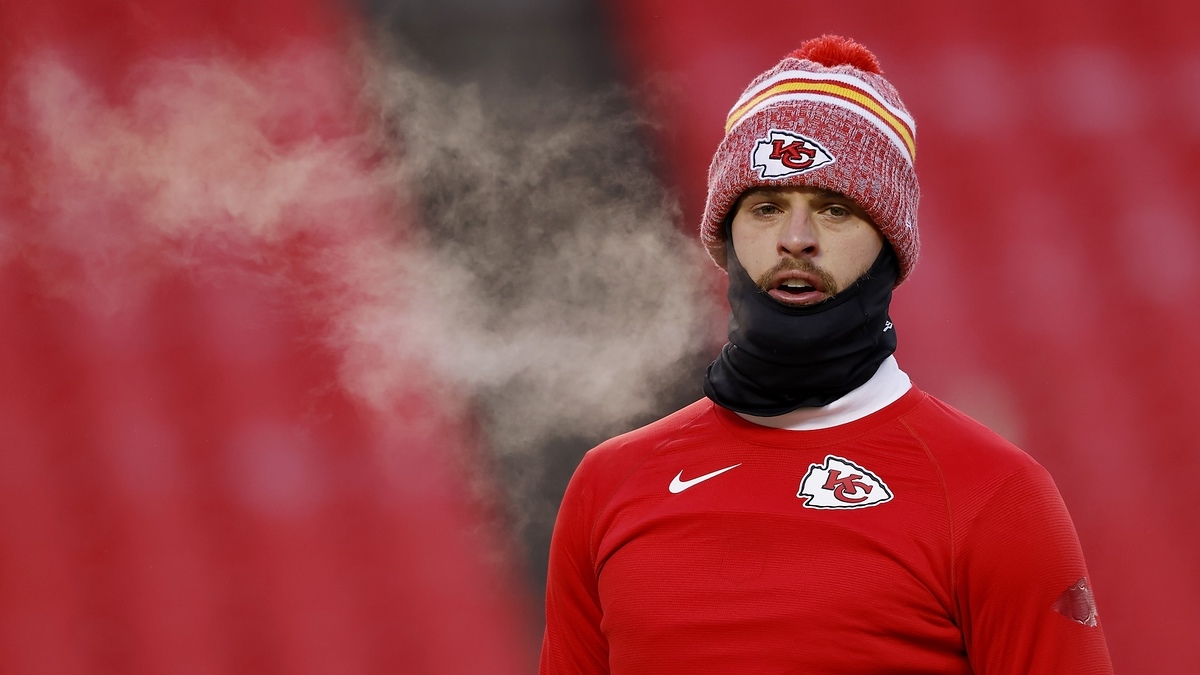 Patrick Mahomes stands on the field, wearing a Kansas City Chiefs knit hat and red hoodie with a black neck warmer. His breath is visible in the cold air