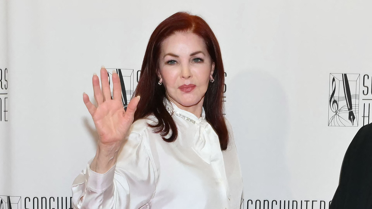 Priscilla Presley is posing on the red carpet at the Songwriters Hall of Fame event, wearing a white blouse and waving to the camera