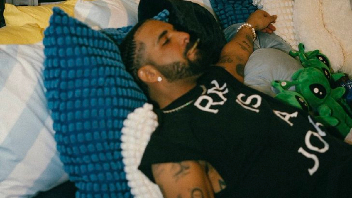 Man wearing a black sleeveless shirt with text "Rel is a joke." He is relaxing on a bed surrounded by pillows and green stuffed toys