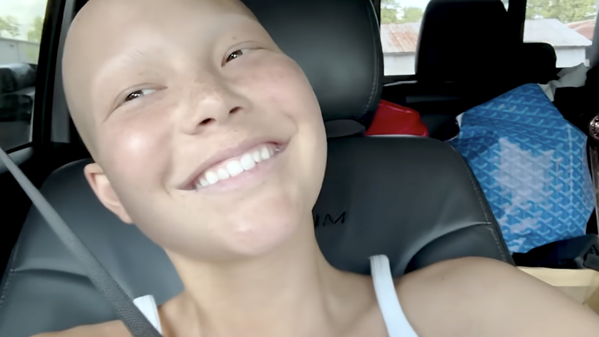 A smiling woman with a shaved head is sitting in a car. She is wearing a white top and appears to be happy. The backseat is filled with bags and various items
