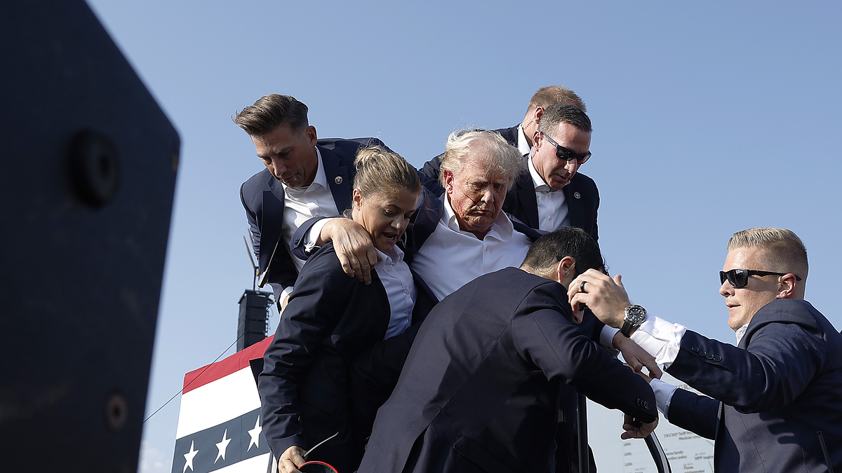 Donald Trump, surrounded by security personnel, being assisted by five men, on a stage with a red, white, and blue backdrop