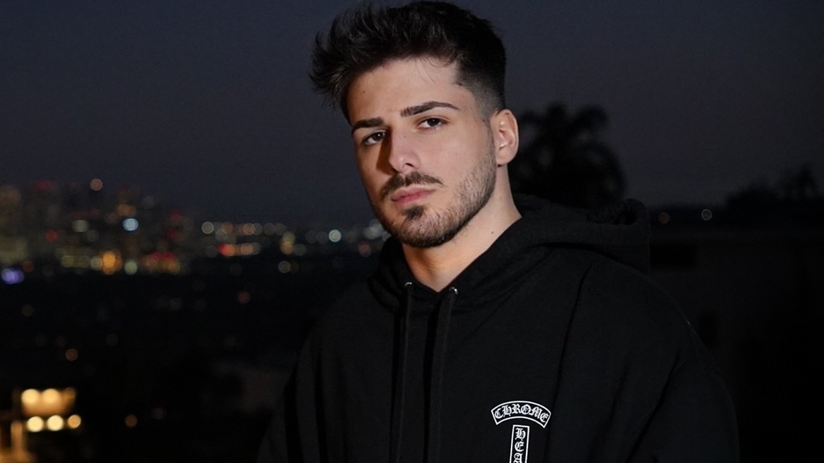 A person stands on a balcony at night with a cityscape in the background. They are wearing a black hoodie