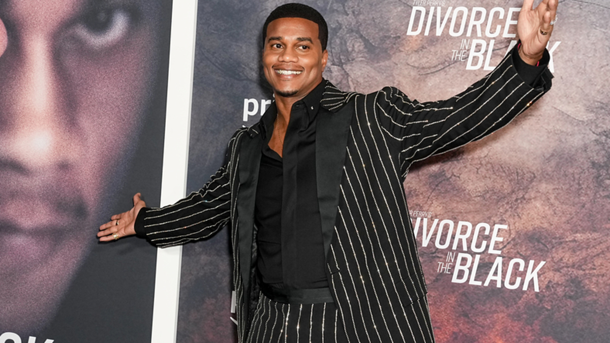 Tyler Perry stands with arms outstretched in front of a "Divorce in the Black" poster at an event, wearing a pinstripe suit