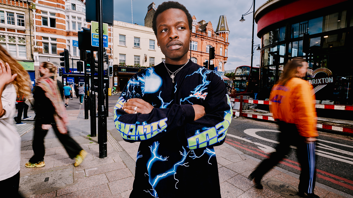 Person on a city street wearing a black shirt with neon blue patterns and graphics. Blurred pedestrians and buildings in the background