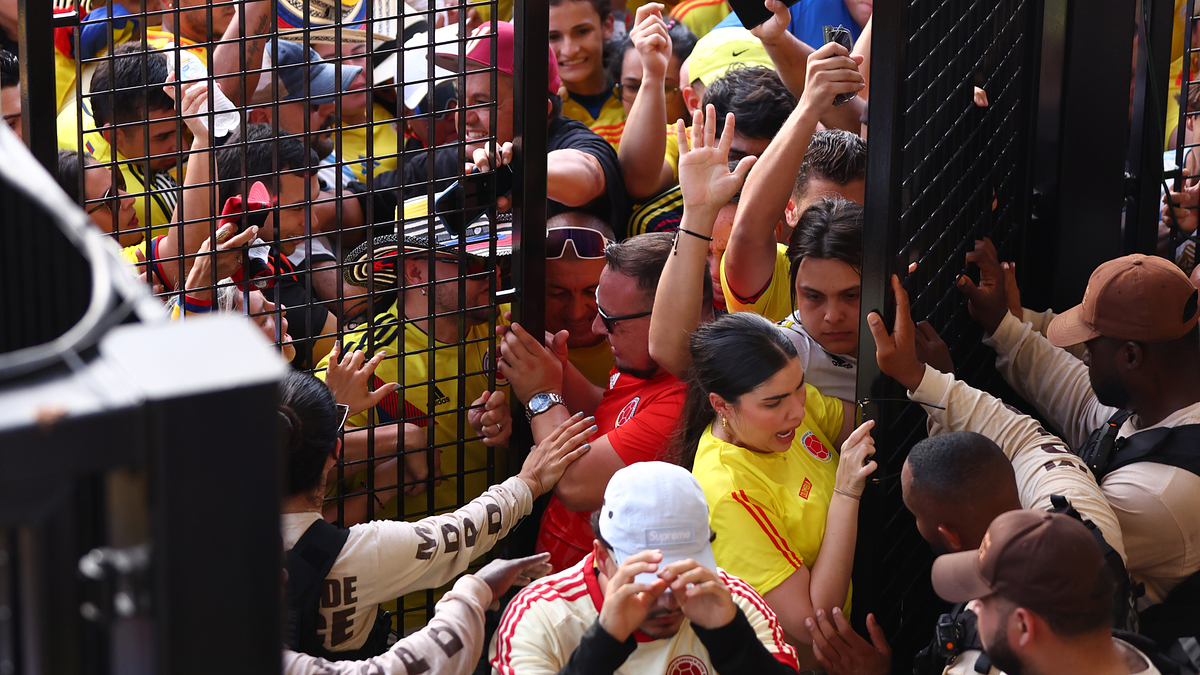 A crowd of sports fans, many wearing yellow jerseys, are entering through a gate with the help of security personnel