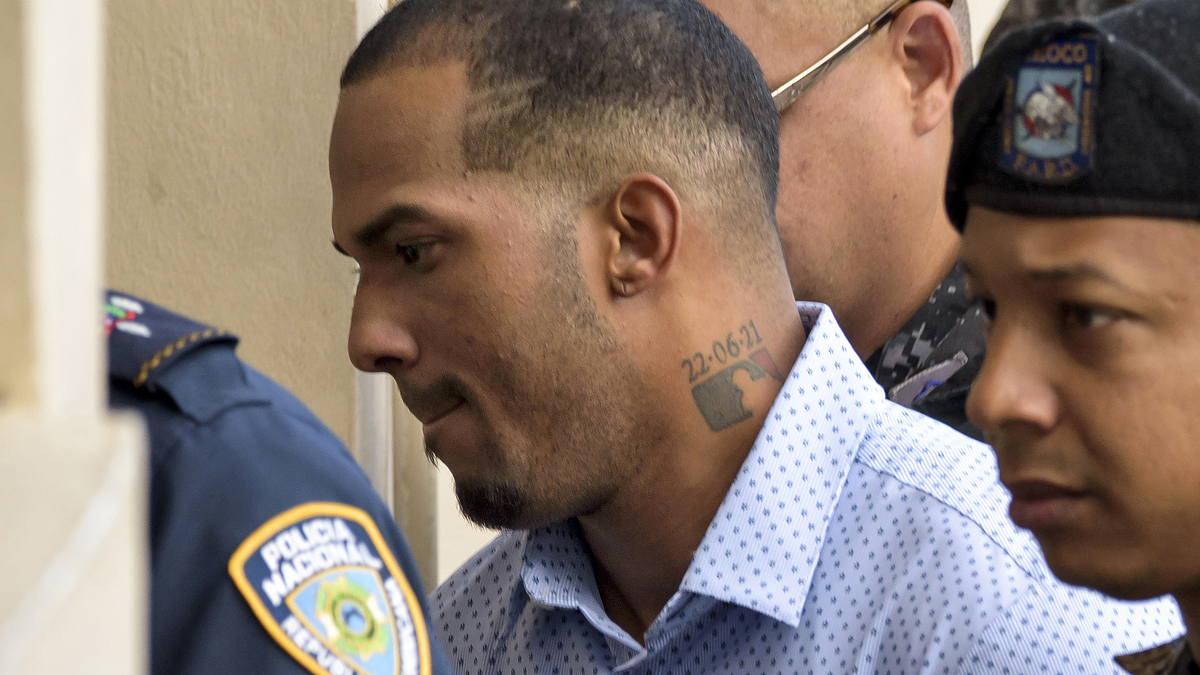 Yankees' pitcher Domingo Germán in a white shirt escorted by police officers, including one in a police beret