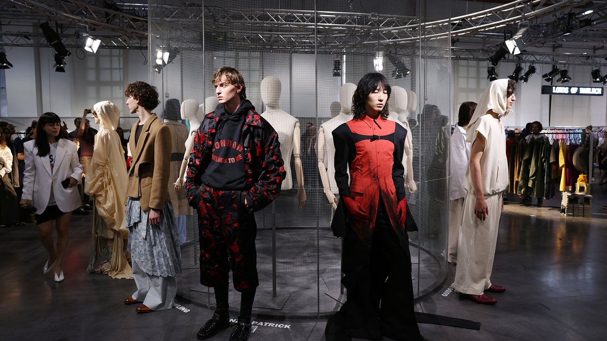 Fashion models on a runway wearing various avant-garde and contemporary outfits. Man at center in red and black patterned clothing. Woman in a stylish red and black outfit