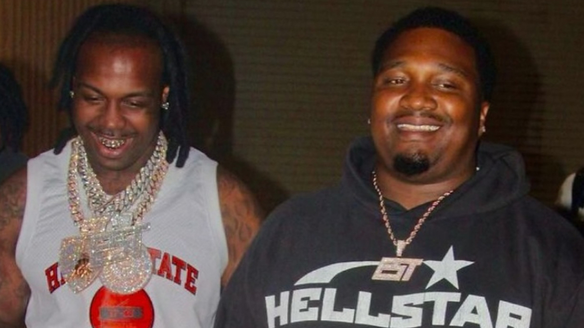 Two men, one wearing a basketball jersey with multiple chains, the other in a hoodie. Both are smiling. Names of persons not provided
