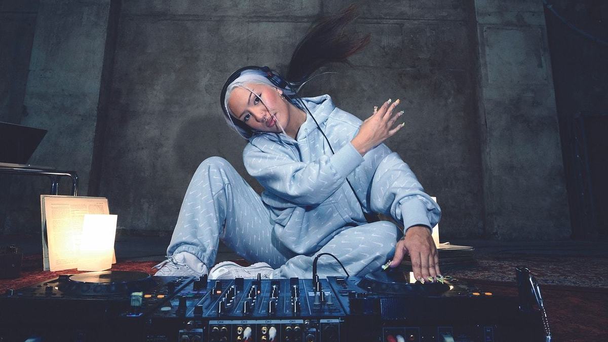Woman wearing a hoodie and sweatpants sits on the floor in a music studio, surrounded by electronic music equipment