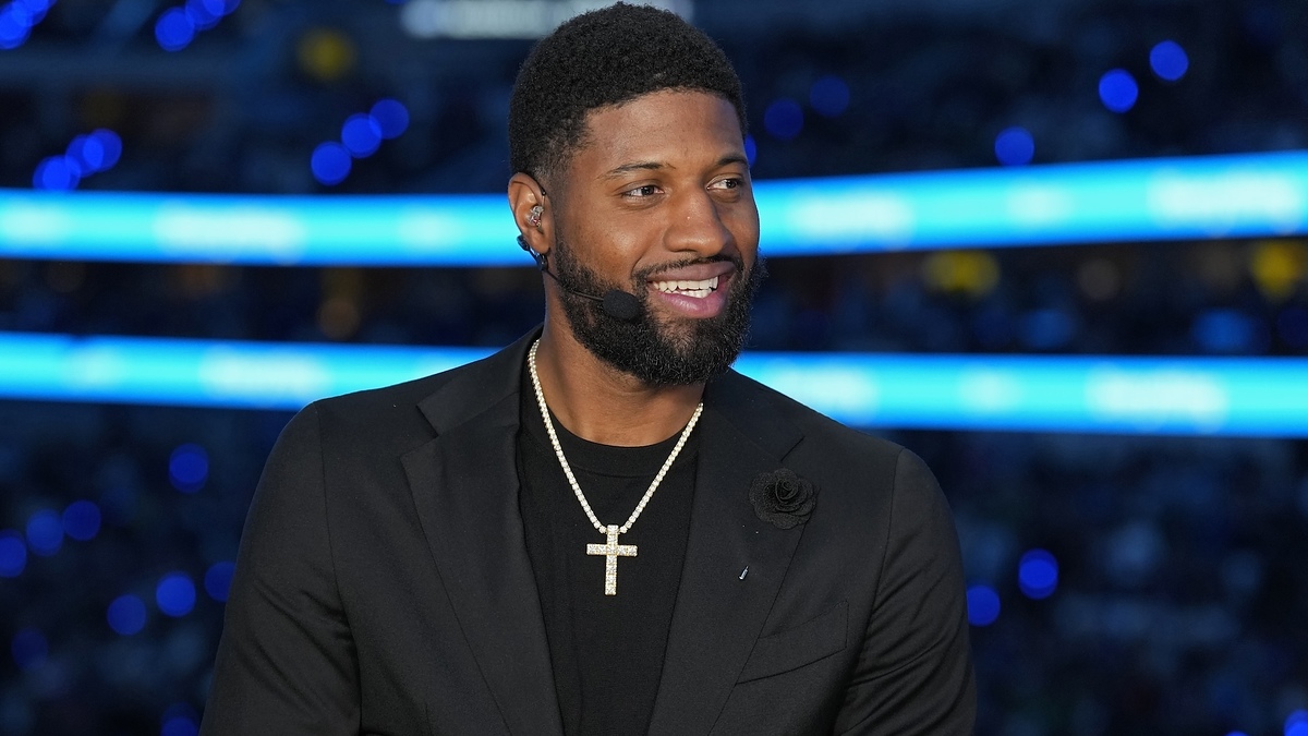 Paul George is smiling while wearing a black suit with a black shirt underneath and a cross necklace. He has a rose pin on his suit