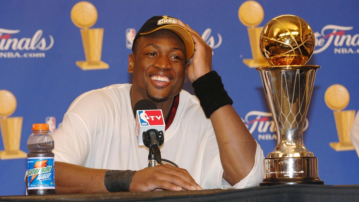 Dwyane Wade smiling, wearing a Championship hat and white shirt, sitting at a press conference table with a Finals trophy and a Gatorade bottle