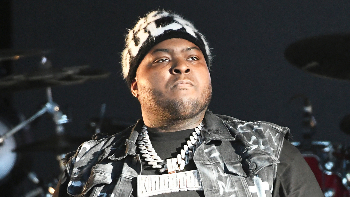 Sean Kingston wearing a patterned hat, black shirt, and a dark denim jacket with chains around his neck, performing on stage