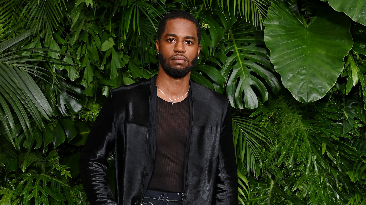 A person stands in front of lush greenery, wearing a black jacket over a sheer black shirt, wide-leg black pants, and a chain accessory