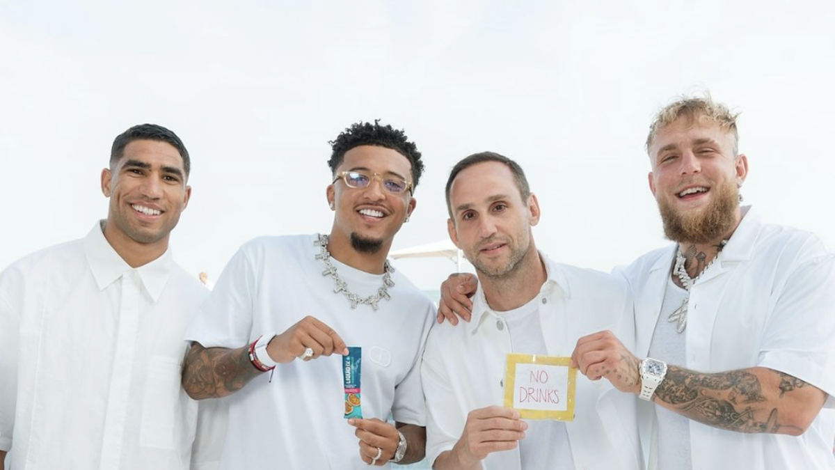 Men holding drinks and laughing together, wearing white outfits. One holds a colorful drink packet, another holds a sign reading "No Drinks."