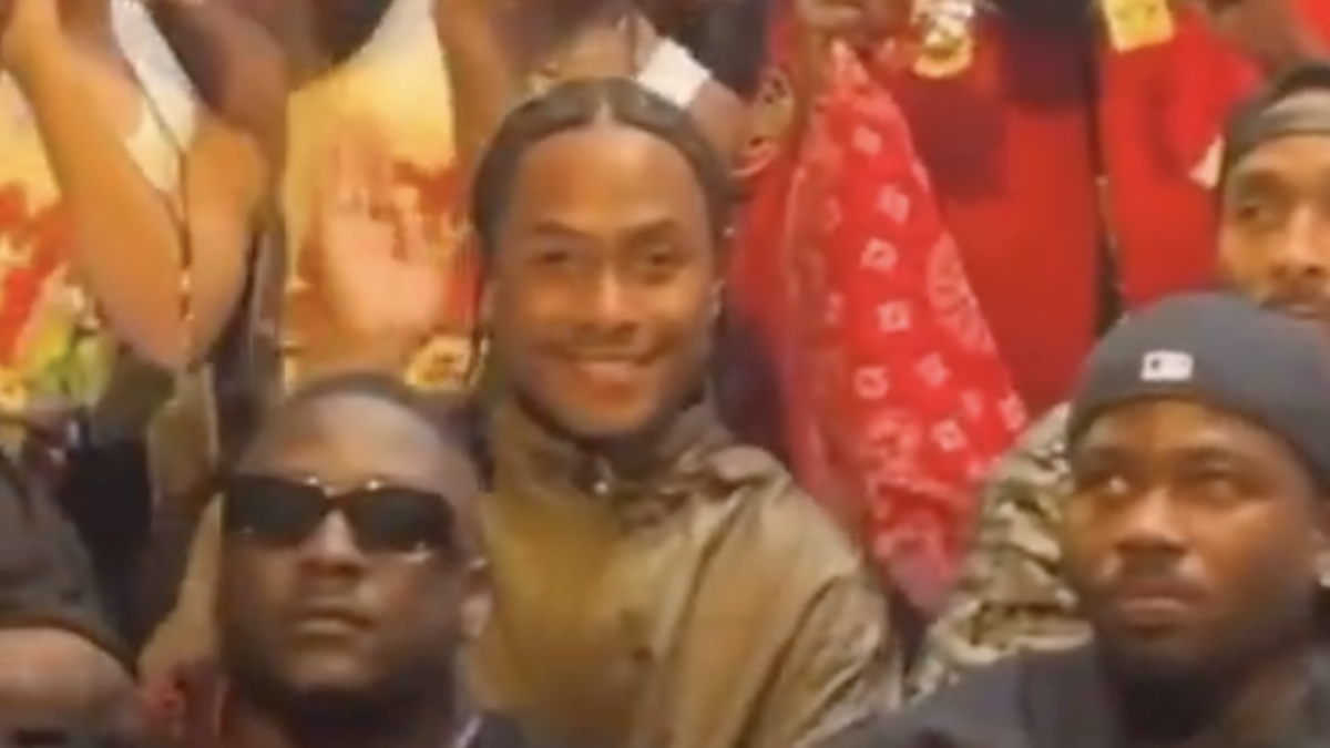 Music group posing together, some making dance moves or gestures, others smiling for the camera. Red cap and gold chains stand out on a person in the back row