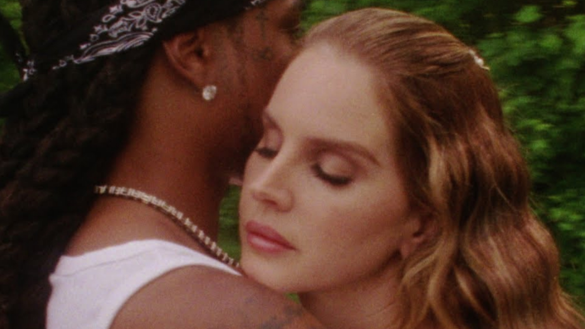Lana Del Rey is seen embracing an unknown man with long braids and a bandana, as they share a tender moment in a forest setting