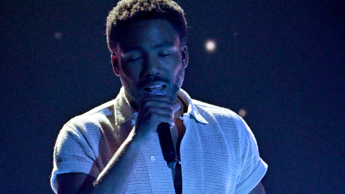 Donald Glover performs on stage, wearing a casual open-neck shirt and holding a microphone against a dark background with scattered lights