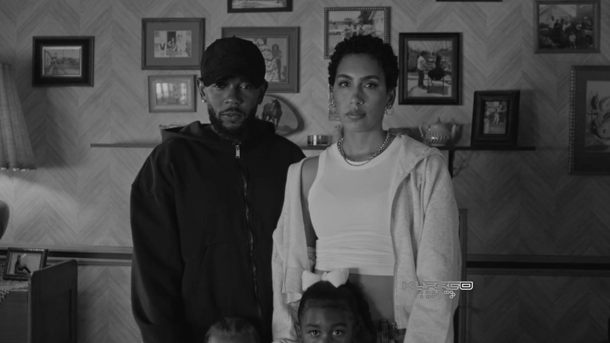 Adults and children standing in front of a wall with framed photos. The man wears a hoodie and cap; the woman wears a sleeveless top and jacket