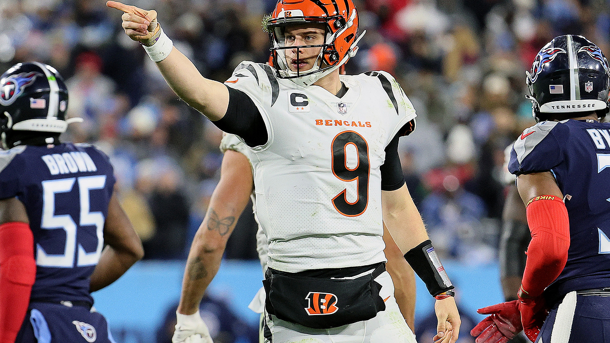 Joe Burrow points on the field during a game while surrounded by opposing players. He wears a Bengals uniform with the number 9
