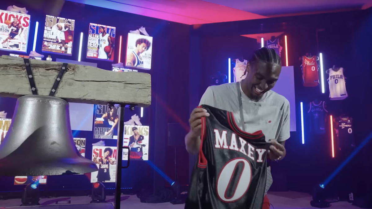 Tyrese Maxey holds his Philadelphia 76ers jersey with a big smile while standing near a large metal bell. Posters with basketball themes decorate the background