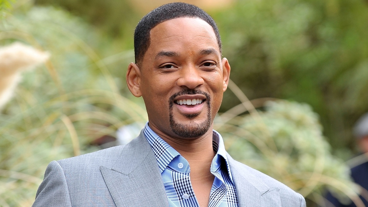 Will Smith smiling in an outdoor setting, wearing a light gray suit with a blue checkered shirt