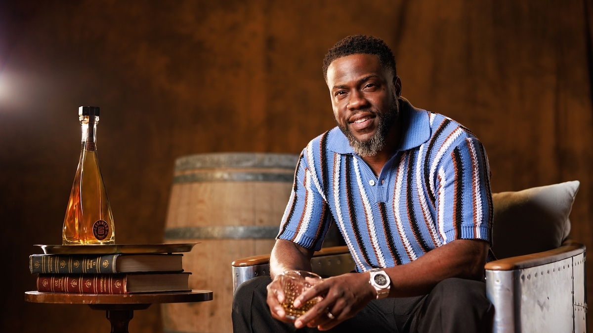 Kevin Hart sits relaxed in an armchair, holding a drink, next to a table with books and a bottle of tequila. He wears a striped polo shirt