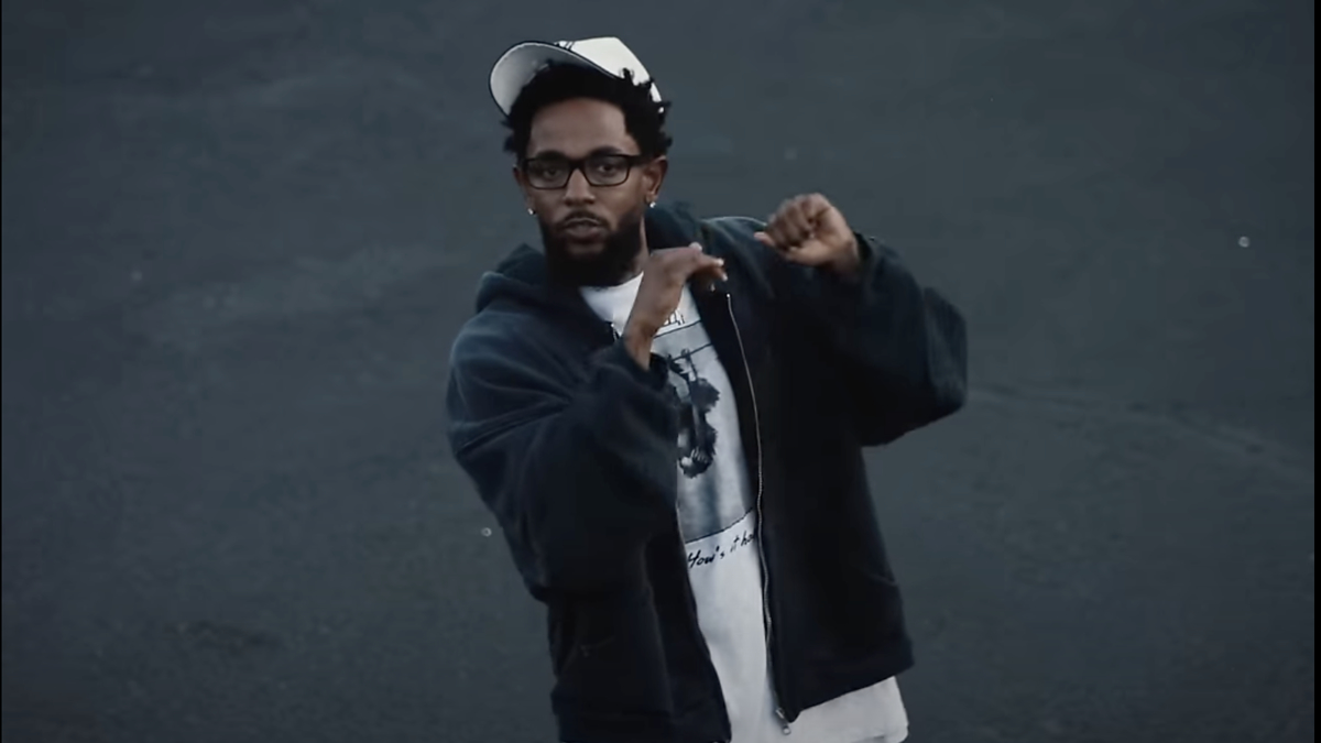 Kendrick Lamar stands outdoors, wearing a dark jacket, graphic tee, and baseball cap, gesturing with his hands