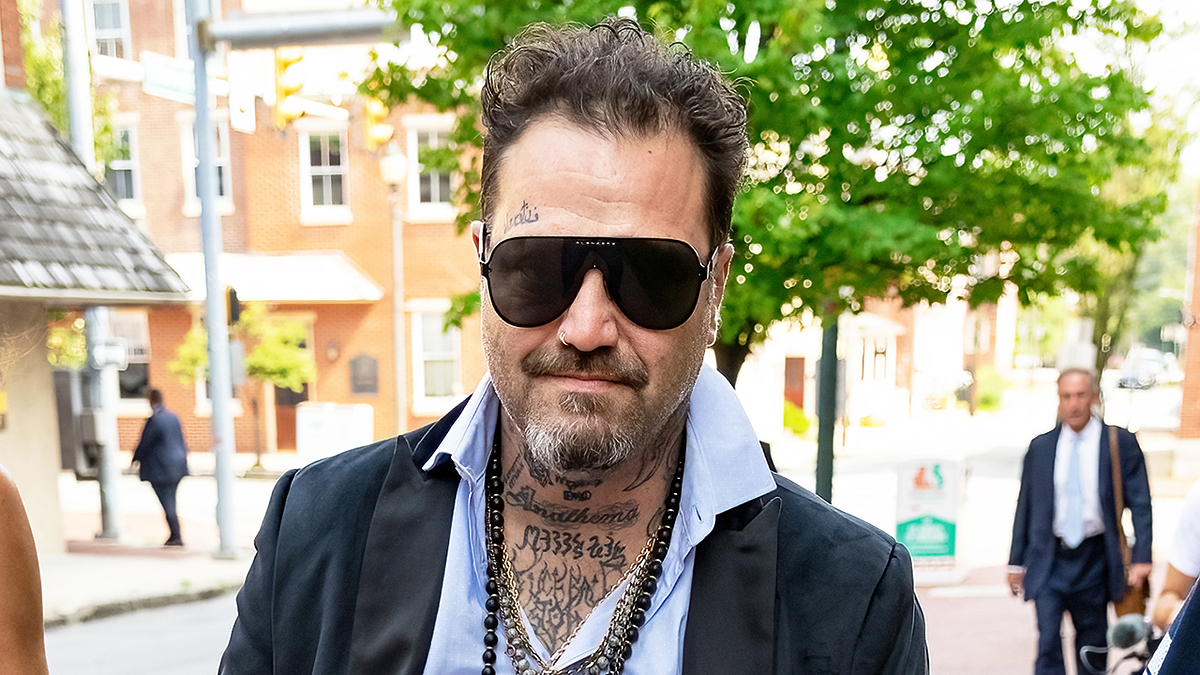 A man with tattoos and a beard, wearing dark sunglasses, a black jacket, a light shirt, and a beaded necklace, walks on a street with buildings in the background