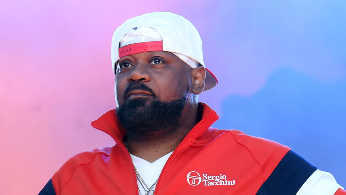 A person wearing a red and white jacket with "Sergio Tacchini" and a white and red backward cap looks into the distance against a blue and purple background